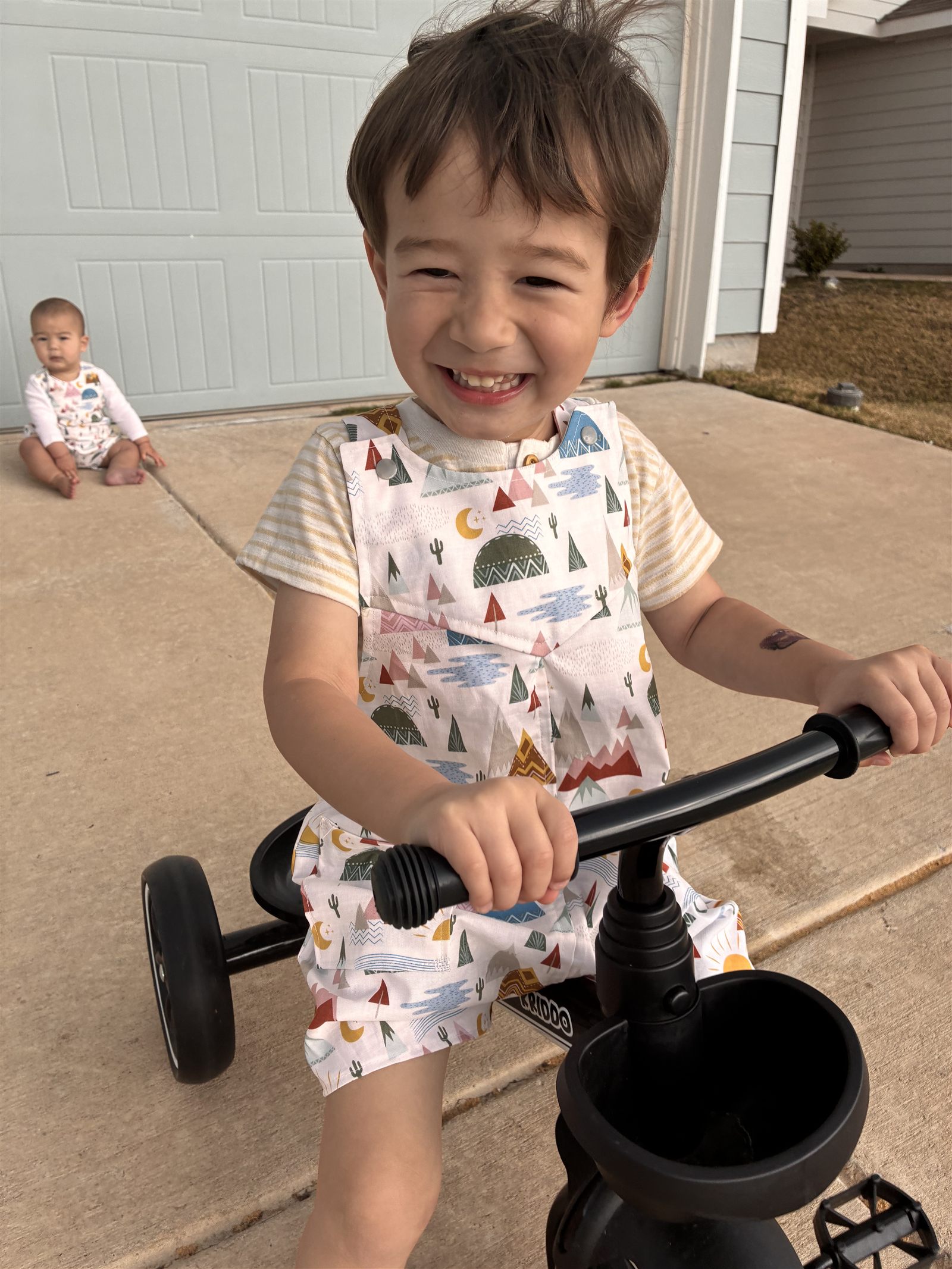 Child on a bike wearing a handmade mountain-print romper with a sibling behind